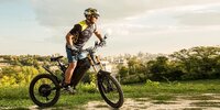 Experienced rider mounts Delfast Top 3.0 electric bicycle on grass with a large city in the backdrop Experienced rider mounts Delfast Top 3.0 electric bicycle on grass with a large city in the backdrop